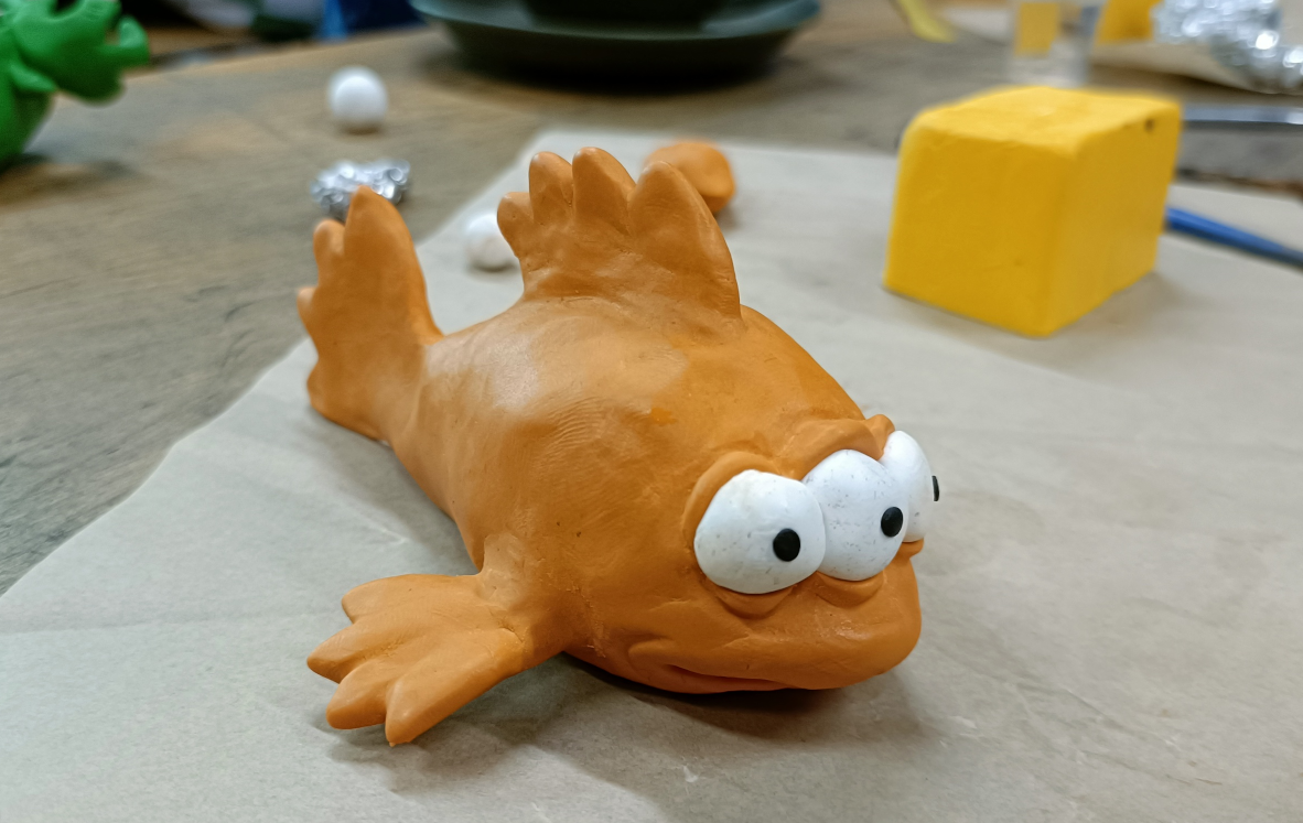 A small handmade polymer clay sculpture of Blinky the fish from the Simpsons, sitting on a bar bench with sculpting tools in the background.