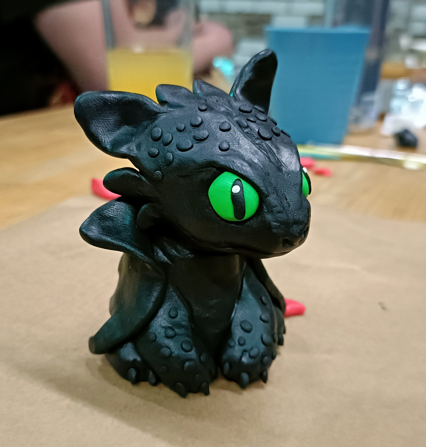 A small handmade polymer clay sculpture of Toothless from How To Train Your Dragon, sitting on a bar bench with sculpting tools in the background.